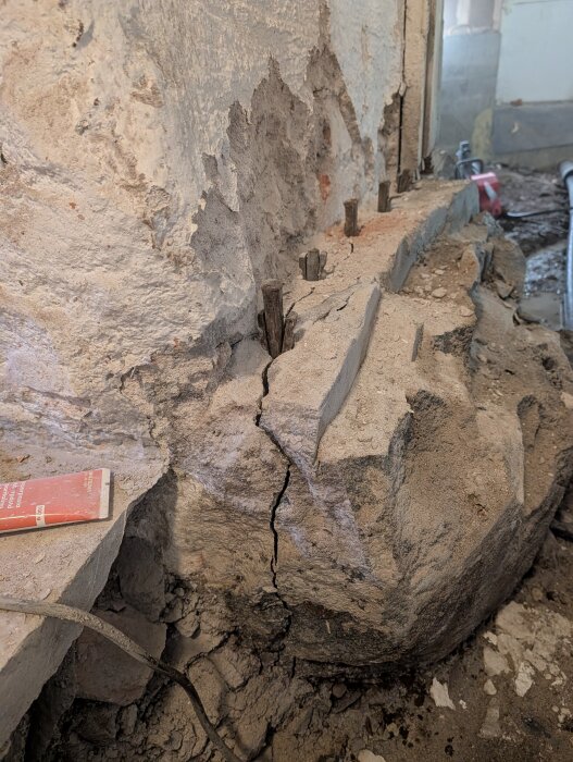Sprucken betongvägg med armeringsjärn synliga och sprickor. Bakomliggande byggarbetsplats med verktyg och kablar i sikte.