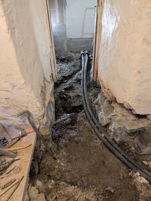 Renoveringsprojekt med grävt smalt schakt i ett hus, exponerade rörledningar och byggverktyg på sidan.