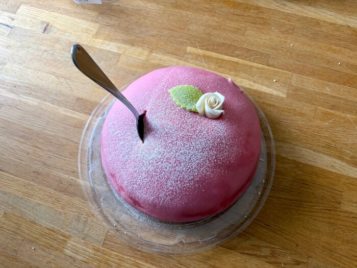 Tårta med rosa marsipan, dekorerad med en vit ros och grönt blad, stående på ett träbord med en sked instucken i toppen.