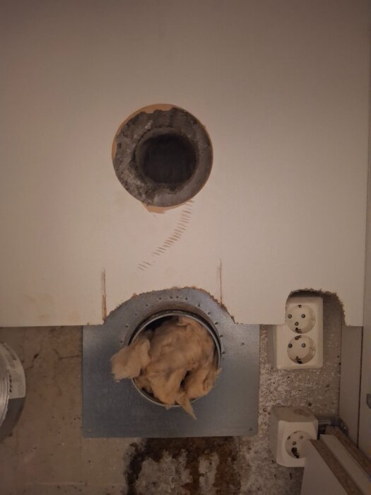 Två runda hål i väggen, det nedre med isolering och en adapter, det övre utan adapter. Cement mellan hålen och eluttag till höger.