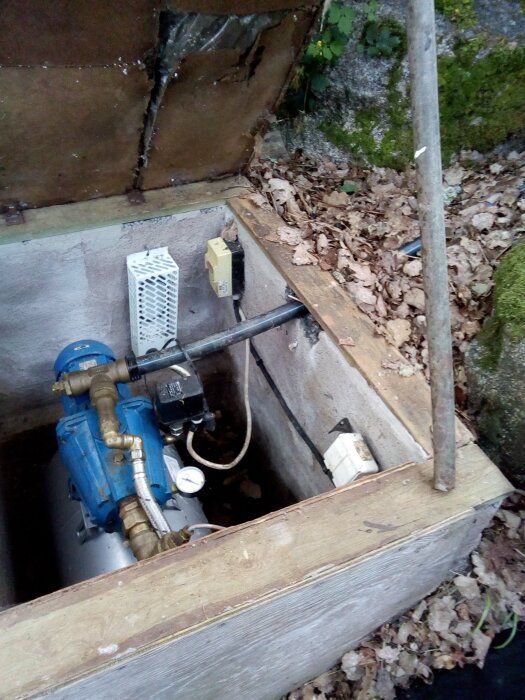 Vattenpumpinstallation i en öppen träbrunn med rör och kablar, omgiven av fallna löv och mossa.