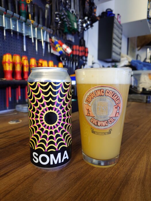 En färgglad burk öl märkt "SOMA" och ett glas öl på en bänk i en verkstad med verktyg i bakgrunden.