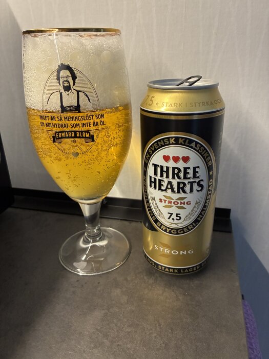 Ett glas med öl och en ölburk av märket Three Hearts, 7,5% alkoholhalt, står på en grå yta. Glaset har ett porträtt och ett citat på sidan.