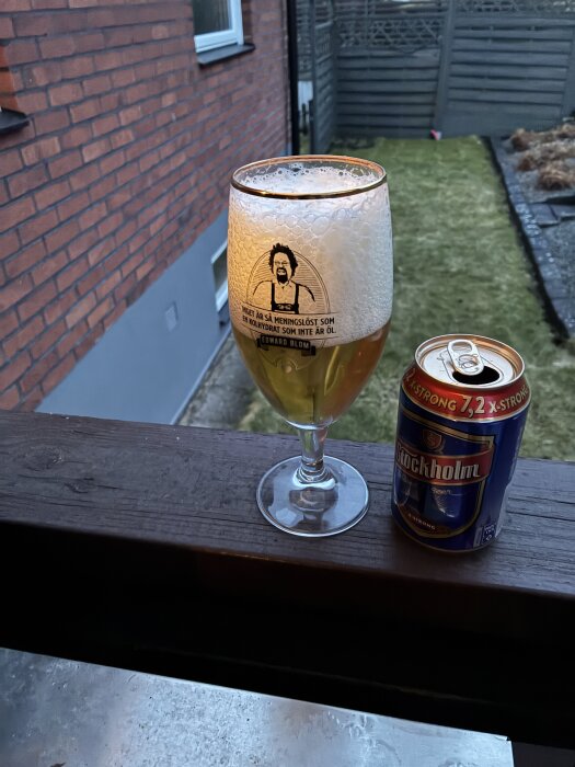 Ett fullt ölglas med skum står bredvid en öppnad Stockholm ölburk på ett räcke utomhus, med en gräsmatta och staket i bakgrunden.