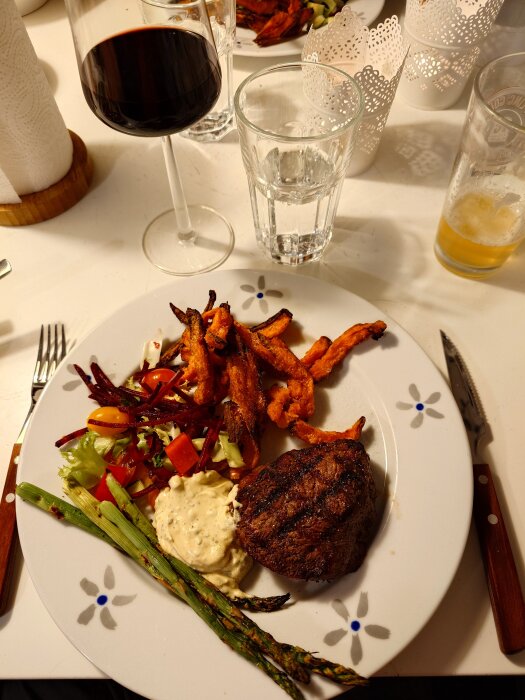 Grillad rostas med sötpotatispommes och grönsaker på en tallrik, rött vin i glas bredvid.
