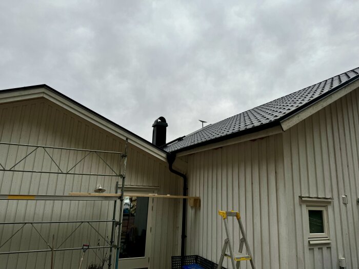 Delvis färdigställd husfasad med takrännor och stuprör, byggställning och stege synliga, himlen molnig.