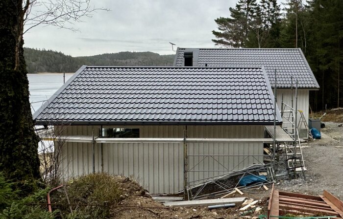 Nybyggd träfasad och tak med mörk plåt på hus, byggställningar runtom och skog i bakgrunden. Utsidan nästan klar.