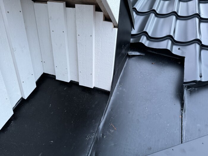 Svart plåttak med detalj av vit träpanel, synlig montering av takavvattning och panelbitar vid ett hörn.