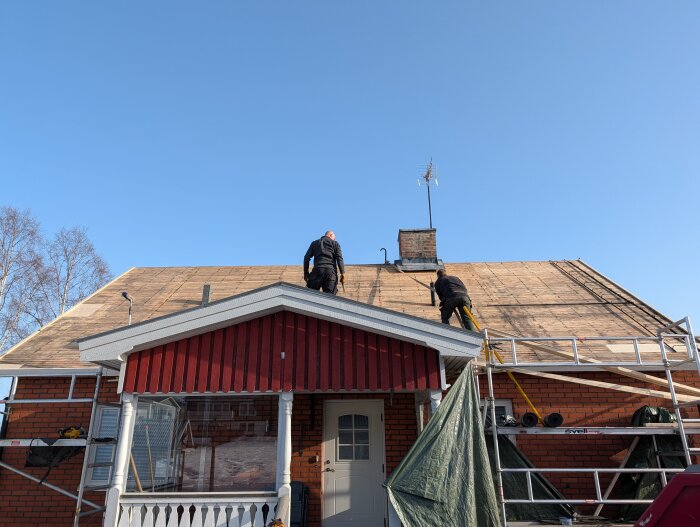 Två personer på taket av ett rött hus arbetar med att byta takpapp. Ställning och stege används för att nå taket under en klar blå himmel.