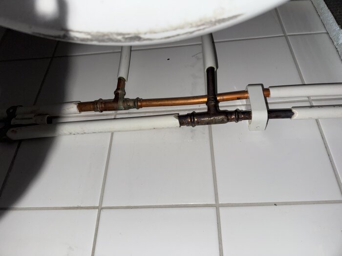 Rörledningar under badkar på vit kakelgolv, koppar- och plastledningar med en koppling i fokus.