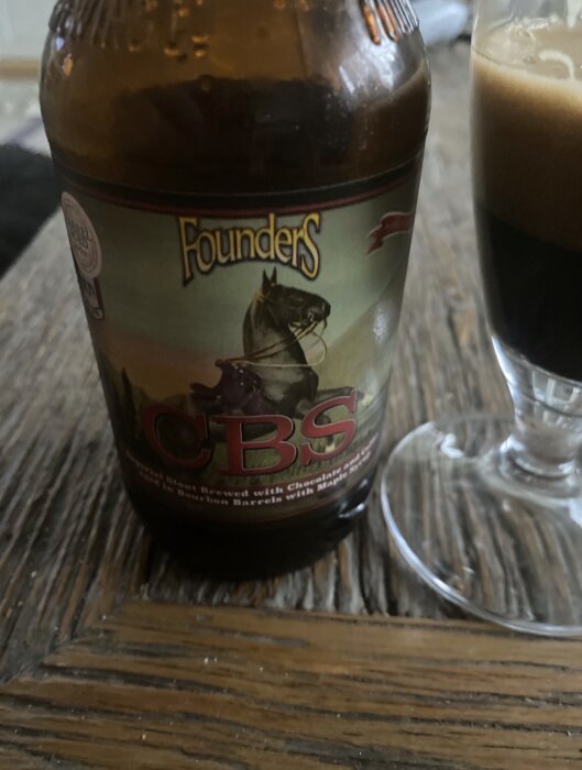 Flaska med etikett "Founders CBS", bredvid ett glas med mörk öl på ett träbord.