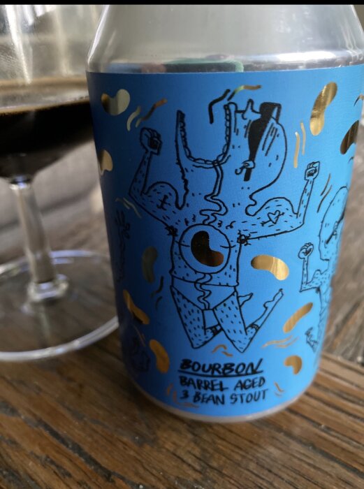 Blå etikett med tecknad figur på en burk "Bourbon Barrel Aged 3 Bean Stout" och ett glas med mörk dryck i bakgrunden.
