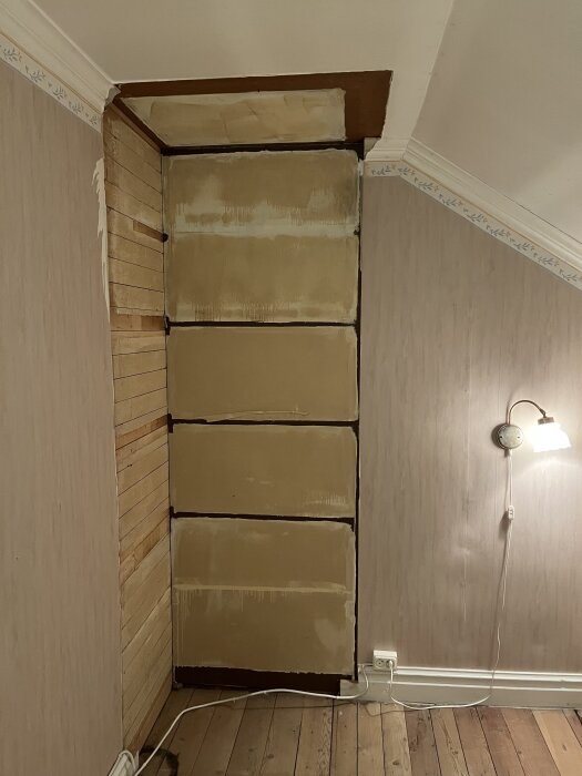 Renovering av vägg i rum med gammal tapet och träpanel, ljus vägglampa och elsladd på trägolv.