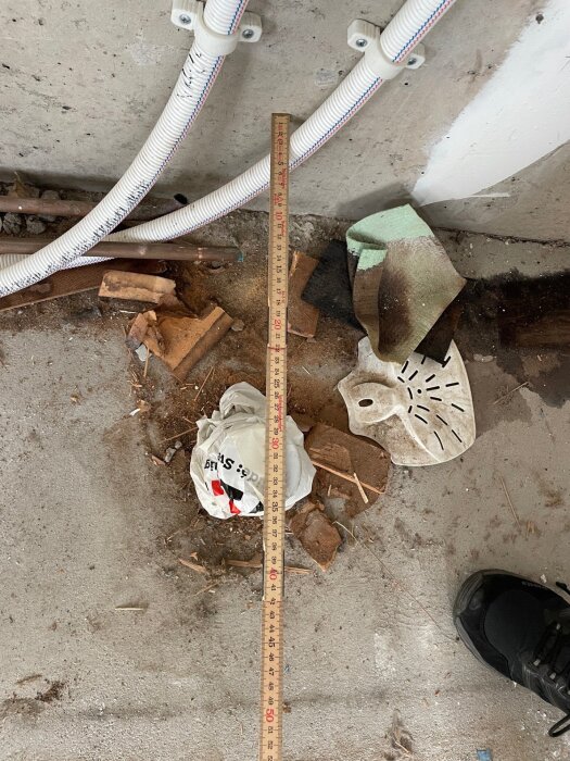 Mätsticka placerad på golvet framför koppar- och plastledningar. En påse och byggmaterialspill är synligt, samt en del av en sko.
