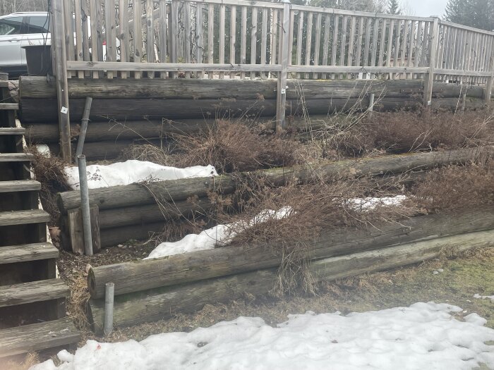 Sluttande tomt med snö och ruttna timmerstockar som fungerar som stödmur, trappen till vänster, med trästaket ovanför.