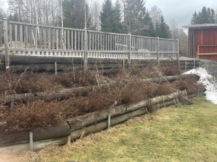 Sluttande tomt med gamla timmerstockar som visar tecken på röta, med ett staket ovanpå och en röd byggnad i bakgrunden.