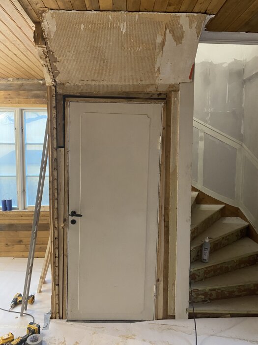 En vit dörr i en hall under renovering, med synligt trävirke och oavslutad vägg. Verktyg och tvålpump syns på golvet.