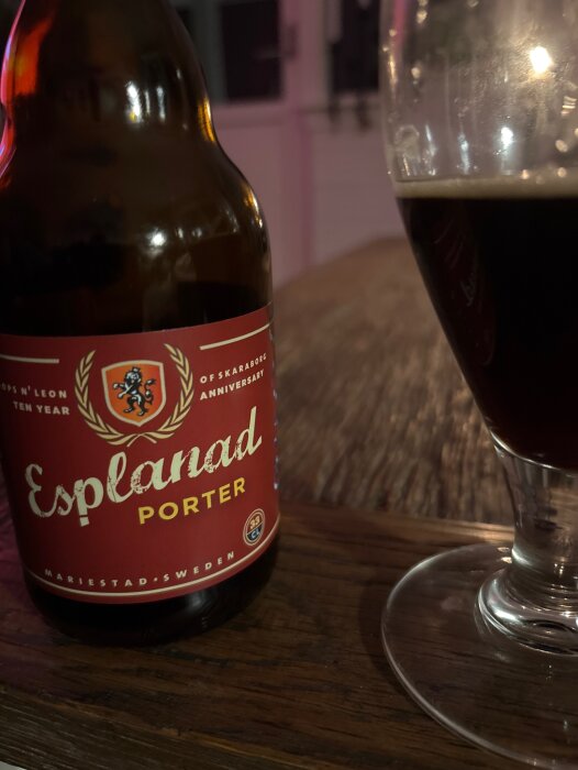 Flaska med Esplanad Porter och ett glas delvis fyllt med porteröl står på ett träbord i ett svagt upplyst rum.