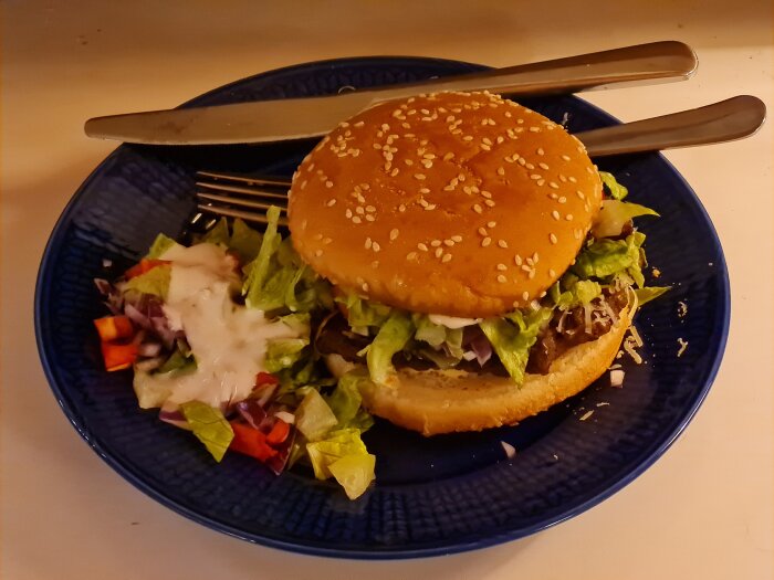 Hamburgare med sallad, riven västerbottensost och dressing på tallrik. Till vänster hackade grönsaker med vitlöksdressing.