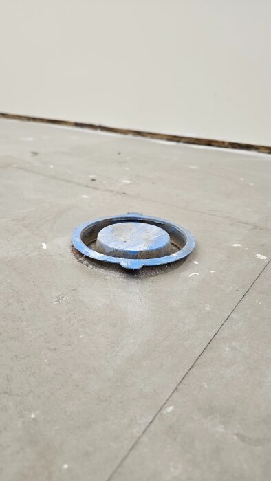 En blå golvbrunnslock installerad i ett cementgolv, omgiven av damm och byggrester.