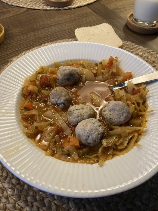 En tallrik kålsoppa med morot, lök, selleri, krossade tomater och frikadeller på ett bord, bröd och ett glas i bakgrunden.