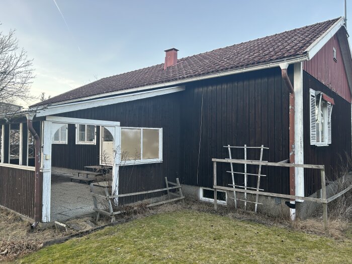 En äldre veranda på ett hus ska rivas, fasaden bakom är fortfarande i gott skick. Kort sida av huset syns med brun träpanel och äldre trästaket.