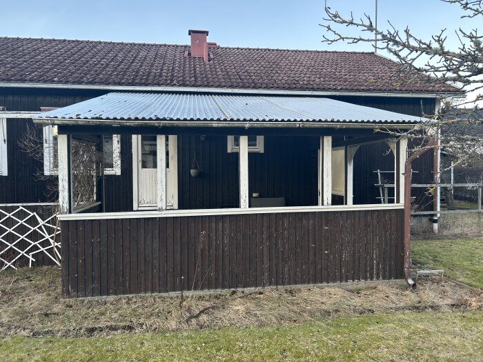 Träveranda med tegeltak på bostadshus, planerad för rivning. Fasad i bakgrunden i relativt gott skick. Torrt gräs och trädgård syns.