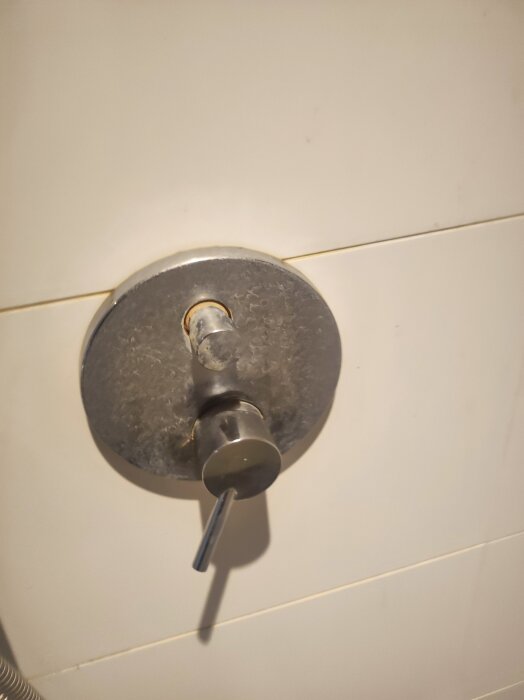 Dold duschblandare med ett vred på vit kakelvägg, visas med ett litet hål under vredet för eventuell demontering.