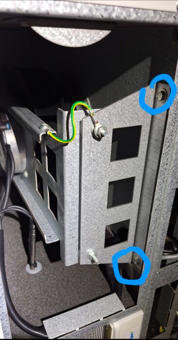 Metallkonstruktion med skruvar och en kopplingsbox, markerade med blå ringar. Grön-gul kabel synlig.