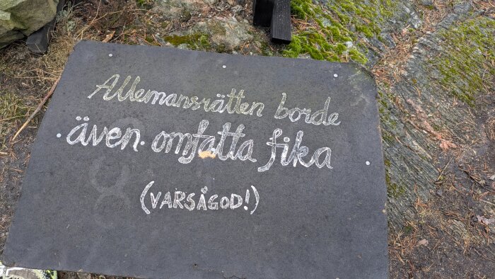 Skylt i naturen med texten "Allemansrätten borde även omfatta fika (Varsågod!)" skrivet med vit färg på en svart bakgrund.