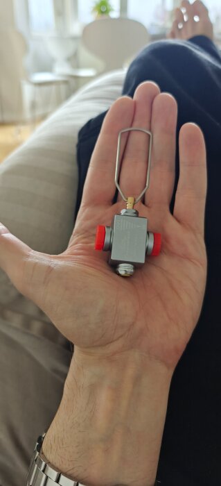 Hand håller adapter för att fylla en mindre primus-behållare från en större, med röd och silverfärgad konstruktion, på en soffa.