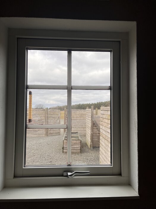 Fönsterruta med röd-målad plastspröjs på väg att lossna. Utsikt över grusad gård med trästaket och himmel i bakgrunden.