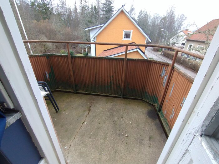 Rostigt balkongräcke och sprucket betonggolv med utsikt över trädgård och bostadshus i bakgrunden.