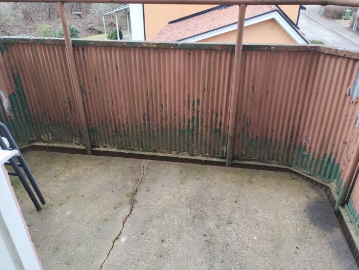 Rostigt balkongräcke och sprucket betonggolv, vy från ovan, omgiven av vegetation och byggnad i bakgrunden.
