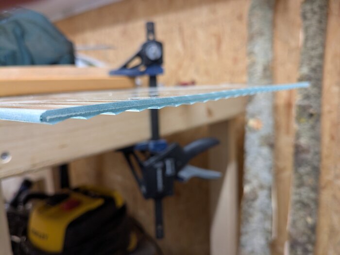 Räfflad glasskiva med grönton och slipade kanter, klämd fast på en arbetsbänk i ett garage, möjlig från ett kasserat vitrinskåp.