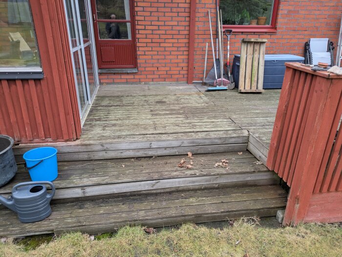 Sliten träaltan med några trappsteg, röd fasad och fönster. Verktyg och ampeldunkar i bakgrunden, blå hink och grön vattenkanna i förgrunden.