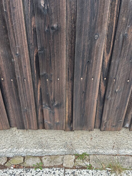 Närbild av träpanel med mörk patina och tecken på slitage. Betonggrund nedanför och viss växtlighet vid kanten.