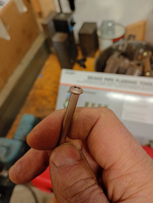 Hand håller ett bearbetat rör skapat med Lidls dubbelkoningsverktyg på en arbetsbänk, maskiner och verktyg i bakgrunden.