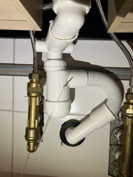 Vattenrör under diskbänk med buntband som fäster plastdelar. Flexibelt teleskoprör och metallkopplingar syns. Används i ett byggprojekt.