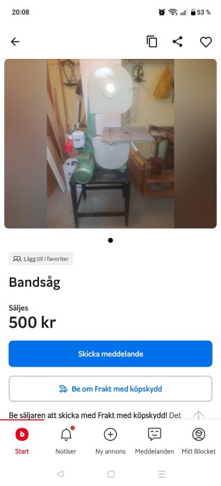 Stationär bandsåg med metallbord och grön motor på svart stativ, omgiven av verktyg och träplankor i garage eller verkstadsmiljö.