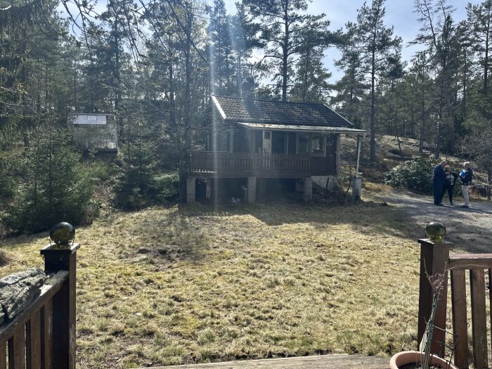 Skogstomt med mossa och ojämn mark framför ett hus, omgiven av träd. Tre personer står på vägen bredvid tomten.