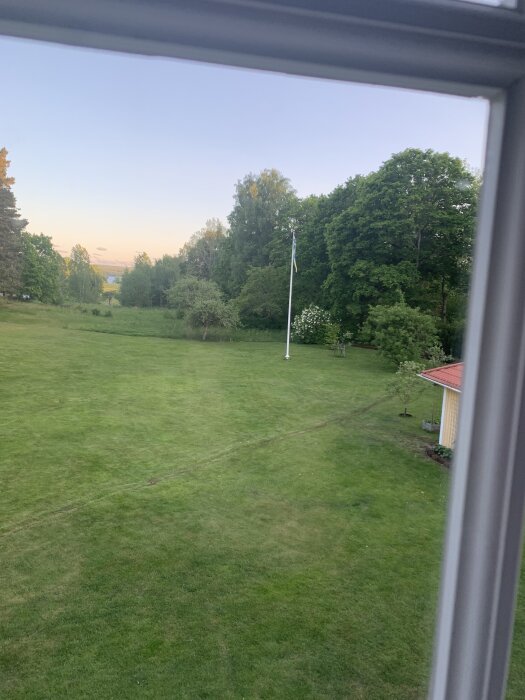 Stor gräsmatta med flaggstång och träd i bakgrunden, sedd från ett fönster.
