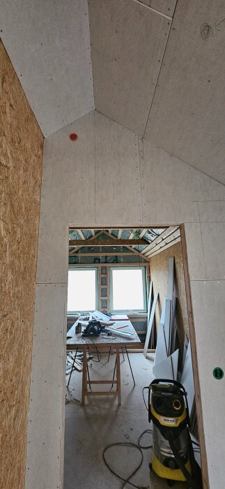 Skivmaterial och snedtak med öppning till rum under renovering, bord med verktyg och dammsugare vid sidan, ljusinsläpp från två fönster.