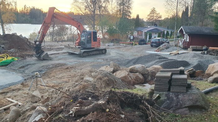 Grävmaskin på en tomt vid en sjö med grushögar och byggmaterial. Skog i bakgrunden och en sommarstuga syns.