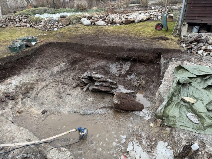 En utgrävd grop i en trädgård med jordiga kanter, isbitar på marken och en pumpslang i vatten. Olika stenar och en presenning syns runt gropen.