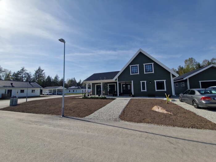 Nybyggt mörkgrönt hus med vita knutar, grusad uppfart och nysått gräs. Bil parkerad bredvid huset och flera grannhus i bakgrunden.