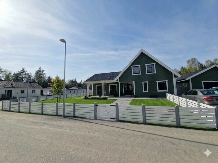 Grått hus med vit staket omgiven av en grön gräsmatta, bredvid två parkeringar med bilar, mot en bakgrund av träd och klarblå himmel.