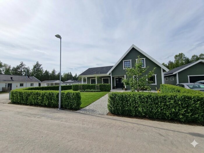 Hus med grå fasad och svart tak omgiven av grön häck och trädgård, med en bil parkerad vid sidan, under en molnig himmel.
