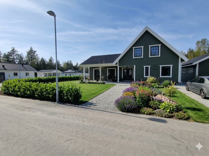 En villa med välskött trädgård inkluderande en rabatt med lagerhägg, andra växter och blommor, omgiven av grön gräsmatta och en stenlagd gång.