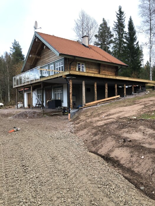 Ett trähus med rött tegeltak, glasräcke och en nybyggd carport, omgiven av träd och grusmark i en lantlig miljö.
